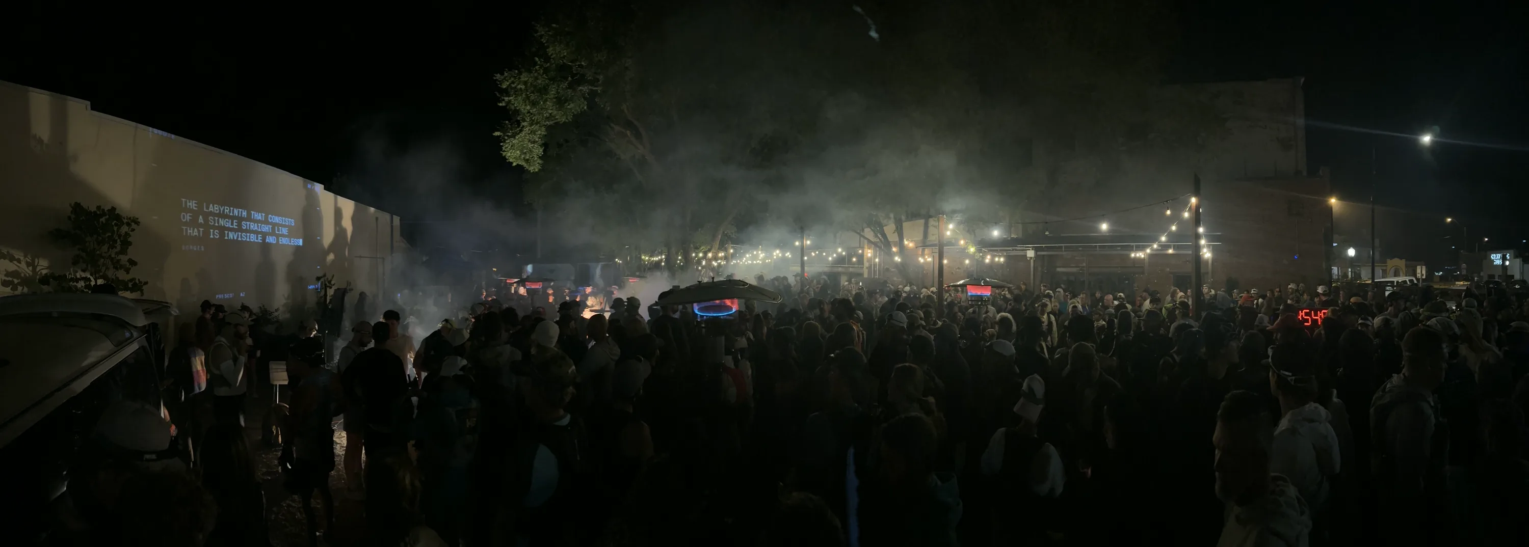 Race night: the wall projecting the Borges labyrinth quote over fog and a crowd of runners under string lights, the race clock glowing red at the far end